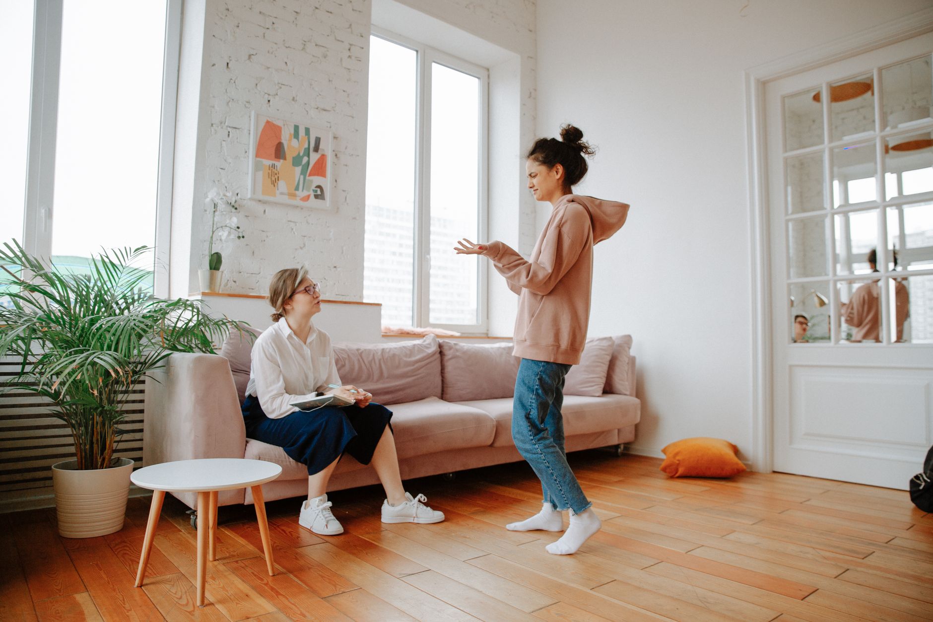 Twee vrouwen in gesprek in een lichte woonkamer met planten.
